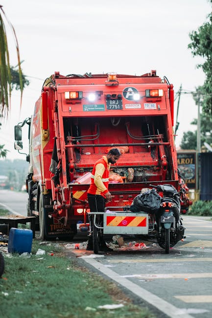 A waste management worker wearing a red high-visibility vest is standing next to a large red rubbish collection truck on a paved road. The truck's rear compaction mechanism is open, revealing debris a