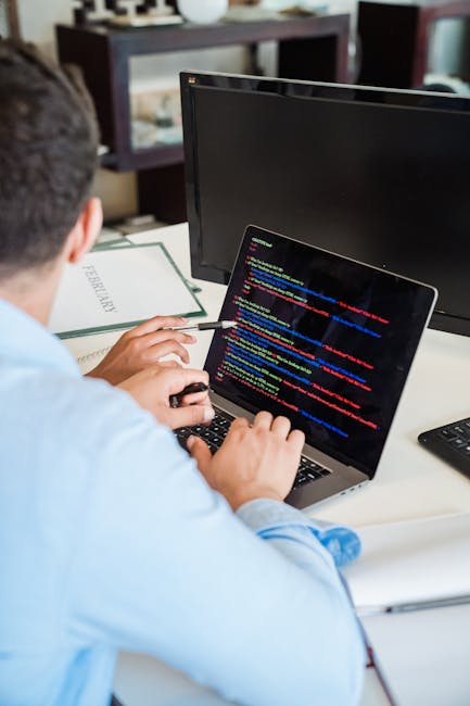 A person wearing a light blue shirt is seated at a white desk, using a laptop computer with a dark code editor displayed on the screen, featuring multicolored lines of programming code. The individual