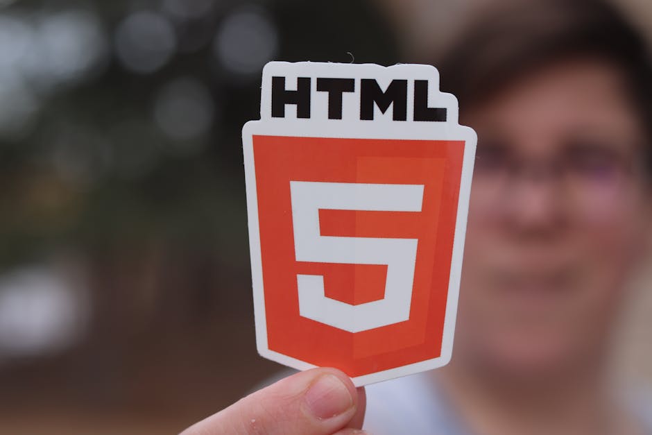 A person wearing a light blue shirt is seated at a white desk, using a laptop computer with a dark code editor displayed on the screen, featuring multicolored lines of programming code. The individual