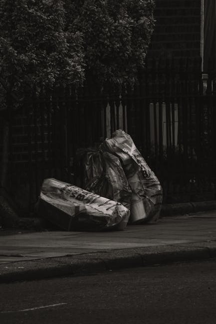 A group of black plastic trash bags, filled and tied at the top, are positioned on the pavement beside a black metal fence. The bags appear to contain household or garden waste and are set against a b
