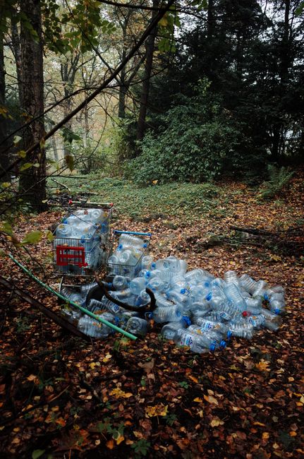 A group of black plastic trash bags, filled and tied at the top, are positioned on the pavement beside a black metal fence. The bags appear to contain household or garden waste and are set against a b